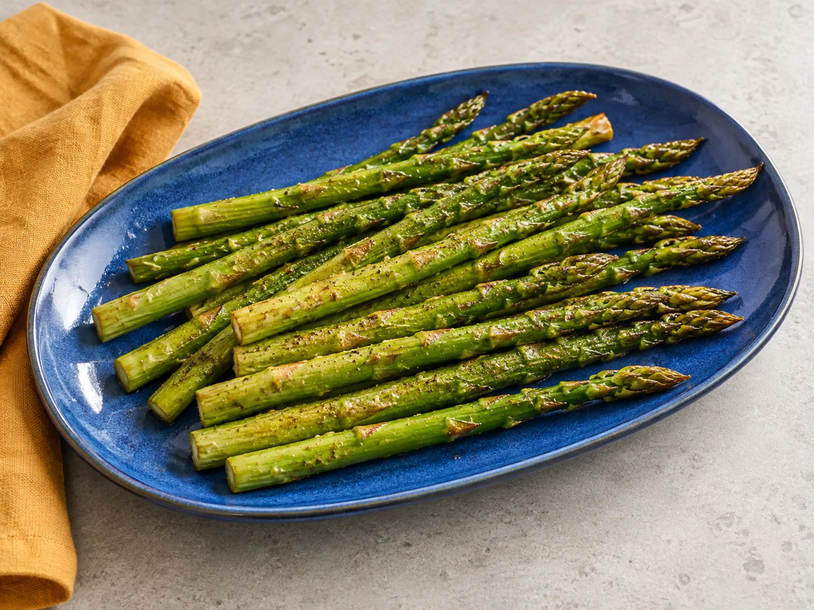 Lightly browned green asparagus spears on a cobalt-blue platter over pale gray stone with amber linen